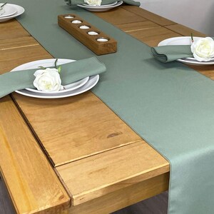 May include: A sage green table runner with a wooden candle holder on a light wood table. The table runner is set with white plates and napkins, and there are white flowers on the table.