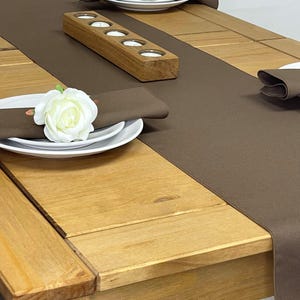 May include: A brown table runner with a wooden candle holder and white plates with white roses on top. The table runner is on a wooden table.