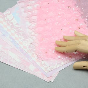 May include: A selection of pink patterned papers with diverse designs, such as floral, lace, and geometric patterns. The papers are fanned out, displaying the different textures and shades of pink. A wooden hand is holding the papers.