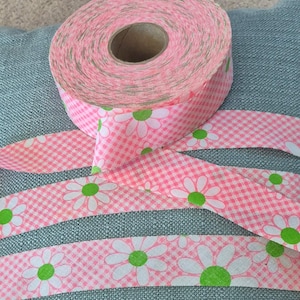 May include: A roll of pink and white gingham ribbon with a daisy pattern. The ribbon is unrolled in strips, revealing the daisy design with white petals and green centers. The ribbon is on a blue-green surface.