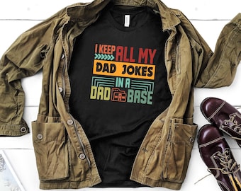 May include: A black t-shirt with a retro-style graphic that says "I keep all my dad jokes in a dad-a-base". The shirt is folded and laid on top of a brown jacket.
