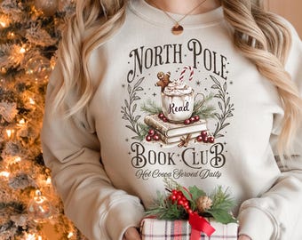 Puede incluir: Una sudadera beige con un diseño navideño que presenta una taza de chocolate caliente, galletas de jengibre y libros. El texto dice "North Pole Book Club Hot Cocoa Served Daily".