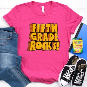 May include: A bright pink t-shirt with the text "FIFTH GRADE ROCKS!" in yellow and orange. The image includes blue jeans, black sneakers, a blue folder, and a box of crayons, suggesting a back-to-school theme.
