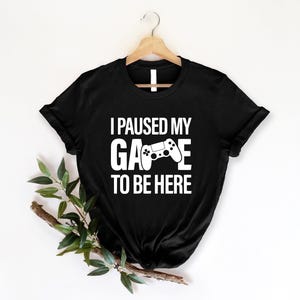 May include: Black t-shirt with the text "I PAUSED MY GAME TO BE HERE" in white. A white graphic of a video game controller is in the center of the word "GAME". The t-shirt is hanging on a wooden hanger.