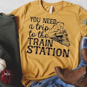 Peut inclure: Un t-shirt jaune moutarde avec du texte noir qui dit "Vous avez besoin d'un voyage à la gare". Une illustration en noir et blanc d'un train avec de la fumée est au-dessus du texte.