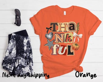 May include: Orange t-shirt with the word "THANKFUL" in a collage style design. The design includes a pumpkin, bow, heart, and star. Black sunglasses and floral print trousers are also visible.