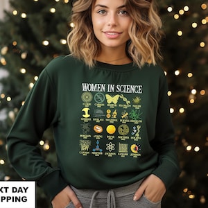 May include: Dark green sweatshirt with the text "WOMEN IN SCIENCE" and illustrations of scientific concepts and women scientists. The sweatshirt features a crew neck and long sleeves. The text "NEXT DAY SHIPPING" is at the bottom.