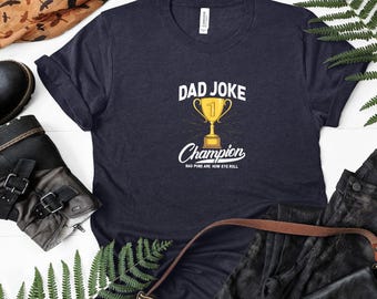 Op de afbeelding: Een marineblauw t-shirt met een witte afbeelding van een gouden trofee met de tekst "Dad Joke Champion" en de woorden "Bad puns are how eye roll".