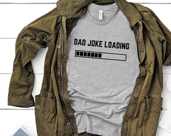 Op de afbeelding: Een grijs T-shirt met de tekst "DAD JOKE LOADING" en een voortgangsbalk eronder. Het shirt is gevouwen en ligt op een bruin jack.
