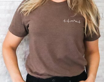 Könnte beinhalten: Ein braunes T-Shirt mit einem weißen Bergketten-Design, das einem Herzschlag ähnelt.