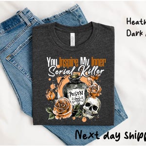 May include: Heather dark grey t-shirt with the text "You Inspire My Inner Serial Killer". The graphic includes a poison bottle, roses, and a skull. The shirt is laid on top of blue jeans.