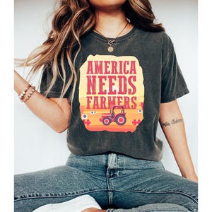 May include: A dark gray t-shirt with a graphic print of a tractor in a field. The text "America Needs Farmers" is printed in red and yellow on the shirt.