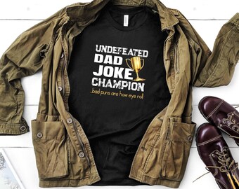 Op de afbeelding: Een zwart T-shirt met witte tekst die "Undefeated Dad Joke Champion" en "Bad puns are how eye roll" luidt. Boven de tekst is een gouden trofee afgebeeld. Het T-shirt is gevouwen en ligt op een bruin jack.