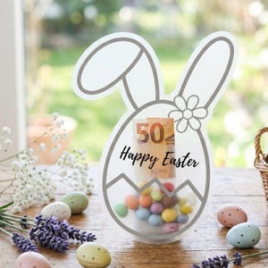 Può includere: Contenitore trasparente a forma di uovo di Pasqua con orecchie di coniglio e un fiore, pieno di caramelle colorate e una banconota da 50 euro. Le parole "Buona Pasqua" sono scritte sul davanti. Uova di Pasqua e un cestino di uova di cioccolato sul tavolo di legno.