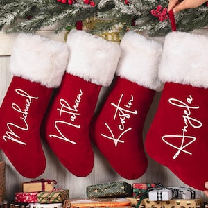 Puede incluir: Cuatro calcetines navideños rojos con ribete de piel sintética blanca y nombres personalizados: Michael, Nathan, Kristin y Angela. Los calcetines cuelgan de una guirnalda con bayas rojas, creando un ambiente festivo.
