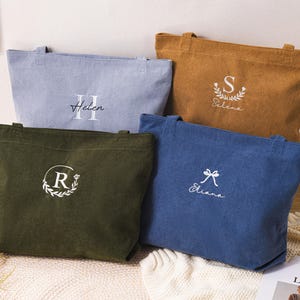 以下が含まれることがあります： ライトブルー、マスタード、オリーブグリーン、ネイビーの4色のコーデュロイのトートバッグ。各バッグには、名前またはイニシャルが白い刺繍でカスタムされています。バッグは長方形で、2つのハンドルが付いています。