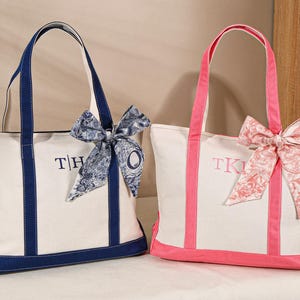 May include: Two canvas tote bags. One is white with navy blue trim, handles, and embroidered initials. The other is white with pink trim, handles, and embroidered initials. Both have decorative bows.