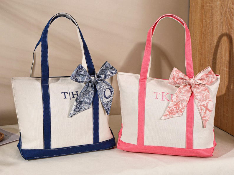 May include: Two canvas tote bags. One is white with navy blue trim, handles, and embroidered initials. The other is white with pink trim, handles, and embroidered initials. Both have decorative bows.