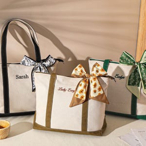 May include: Three canvas tote bags with personalized names and decorative scarves. The bags are off-white with black, green, and brown accents. Each bag has a different scarf design, including floral and patterned options.
