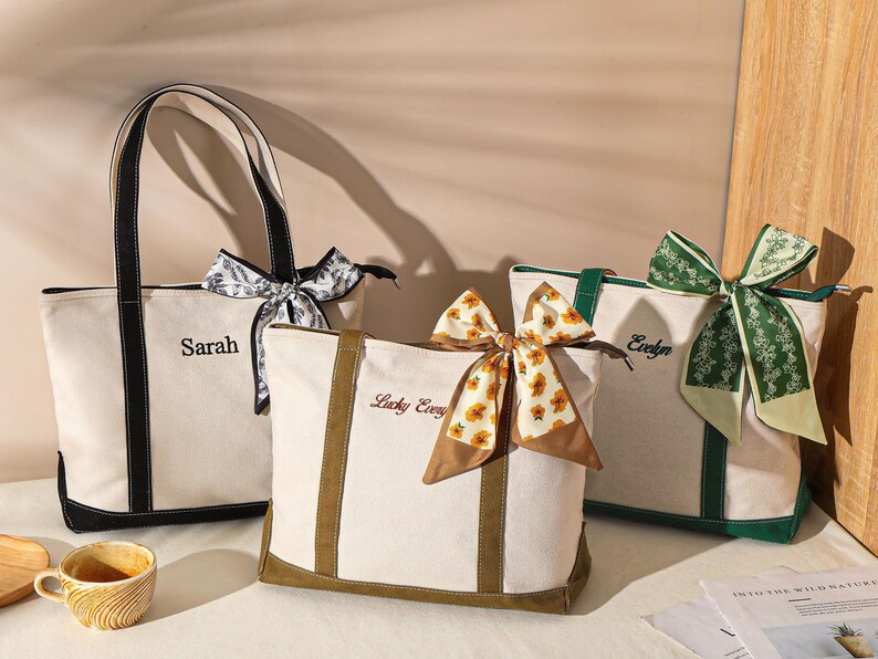 May include: Three canvas tote bags with personalized names and decorative scarves. The bags are off-white with black, green, and brown accents. Each bag has a different scarf design, including floral and patterned options.