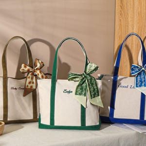 May include: Three canvas tote bags with different colored trim and handles. The bags are cream-colored with brown, green, and blue accents. Each bag has a decorative bow and embroidered text: "Lucky Every", "Evelyn", and "Good Lu".
