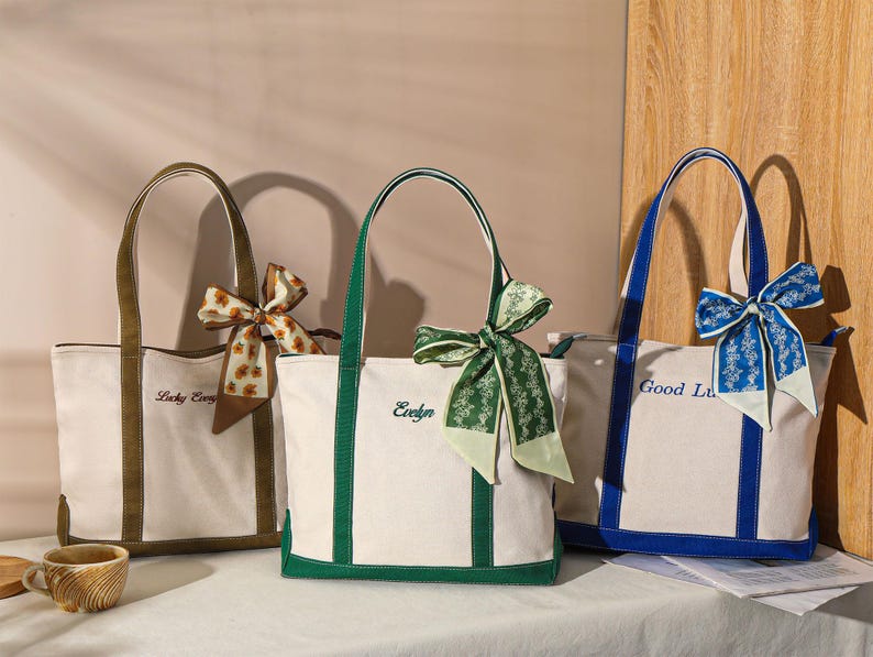 May include: Three canvas tote bags with different colored trim and handles. The bags are cream-colored with brown, green, and blue accents. Each bag has a decorative bow and embroidered text: "Lucky Every", "Evelyn", and "Good Lu".