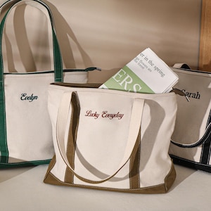 May include: Three canvas tote bags in cream with contrasting trim and handles. One bag has green trim and the name "Evelyn" embroidered. Another has brown trim and "Lucky Everyday" embroidered. The third has black trim and the name "Sarah".