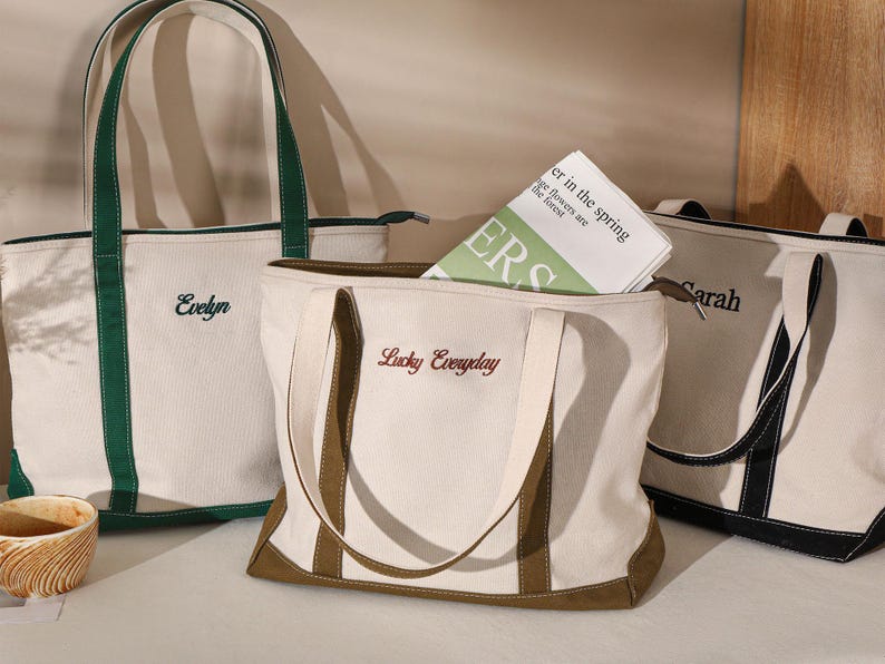 May include: Three canvas tote bags in cream with contrasting trim and handles. One bag has green trim and the name "Evelyn" embroidered. Another has brown trim and "Lucky Everyday" embroidered. The third has black trim and the name "Sarah".
