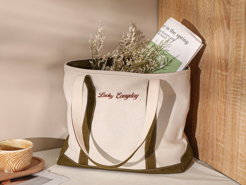 May include: Cream-colored tote bag with olive green accents and handles. The bag features the embroidered text "Lucky Everyday" in a reddish-brown color. The bag is filled with dried flowers and a newspaper with the headline "er in the spring".