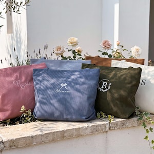 Puede incluir: Una colección de bolsos tote de pana en varios colores, como rosa, azul y verde oliva. Cada bolso presenta bordados personalizados con nombres o iniciales. Los bolsos se exhiben al aire libre con flores en el fondo.