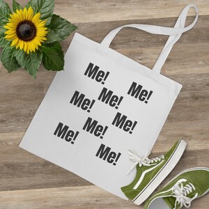Puede incluir: Bolsa de tela blanca con la palabra "Me!" repetida varias veces en texto negro.