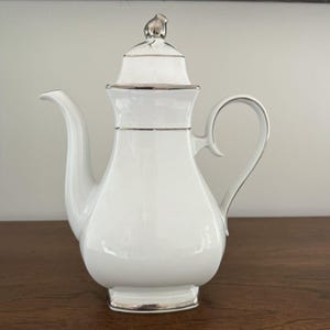 May include: White porcelain coffee pot with a silver trim and a silver knob on the lid.