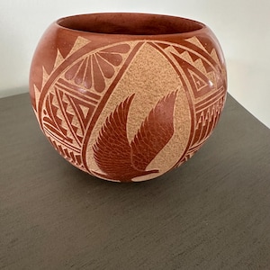 Peut inclure: Un bol rond en céramique de couleur terre cuite avec des motifs gravés. Le bol présente un motif central d'oiseau en vol, entouré de motifs géométriques. Le bol est une pièce décorative, probablement pour l'exposition ou le stockage.