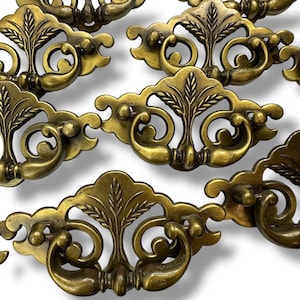 May include: A collection of antique brass drawer pulls. Each pull features an ornate design with a central leaf motif, curved handles, and decorative flourishes. The hardware has a warm, aged patina.
