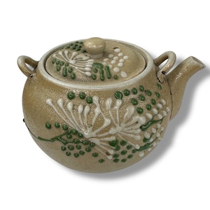 May include: A beige ceramic teapot with a lid, decorated with green and white floral designs. The teapot has a rounded body and a spout on the side.