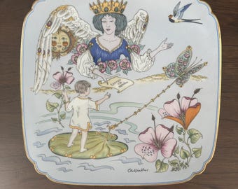 May include: Square decorative plate with a light blue background and a gold trim. The plate features a detailed illustration of an angel, a child, flowers, and a butterfly. The plate has the artist's signature, "Ole Winther".