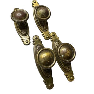 May include: Four antique brass cabinet pulls with a distressed finish. Each pull features a round knob and an elongated backplate with floral detailing. The pulls have a vintage aesthetic, perfect for adding character to furniture.