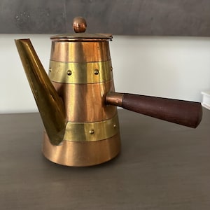 May include: A copper and brass coffee pot with a wooden handle. The pot has a cylindrical body with brass bands and rivets. The spout is a contrasting gold color. The lid is topped with a small brass knob.