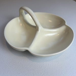 May include: Cream-colored, three-section serving dish with a handle. The dish has a clover-like shape with rounded sections. The handle is arched and connects to the center of the dish. The surface is smooth and glossy.