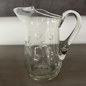 May include: A clear glass pitcher with a decorative etched pattern of circles and lines. The pitcher has a rounded body and a handle.
