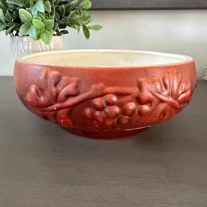 May include: A reddish-brown ceramic bowl with a textured surface and a cream-colored interior. The bowl features a raised relief design of grape clusters and leaves around the exterior. The bowl is a decorative piece for the home.