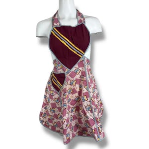 May include: A vintage apron with a halter neck design. The apron features a heart-shaped bodice in burgundy with yellow and blue stripes. The skirt has a pink and white floral pattern with blue trim. The apron is displayed on a white mannequin.