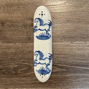 May include: A long, oval-shaped ceramic wall plaque with a glossy cream-colored glaze. The surface features two hand-painted blue horse figures in a prancing pose, each positioned above a small patch of grass. There are two small circular holes.