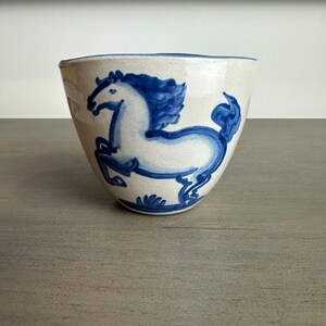 May include: A speckled off-white ceramic bowl featuring a hand-painted blue horse illustration. The horse is depicted in a prancing pose with a flowing mane and tail. The bowl has a wide, open top and a tapered base, with a blue rim accent.