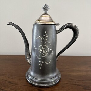 May include: A silver teapot with a floral design on the side. The teapot has a silver lid with a knob on top. The teapot is sitting on a wooden surface.