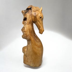 May include: A wooden sculpture of a horse's head and neck, carved from a light brown wood. The sculpture has a natural, rustic appearance with visible wood grain and a rough texture. The horse's head is tilted, and the mane is detailed with carved lines.