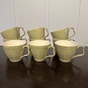May include: A set of eight green ceramic teacups with a textured, vertical striped pattern. The cups have a curved handle and a cream-colored interior. The teacups are arranged on a dark brown surface.