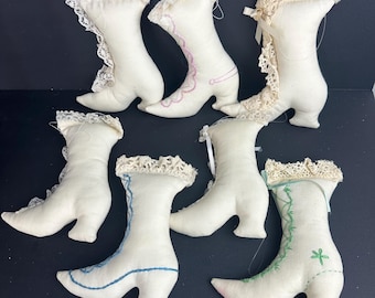 May include: A collection of seven off-white fabric boots with lace trim and embroidered details. Some boots feature pink, blue, or green stitching. The boots have a vintage aesthetic.