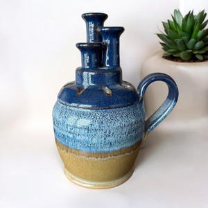 May include: A blue and brown ceramic vase with three openings on top and a handle. The vase has a speckled glaze.