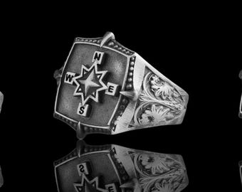 Könnte beinhalten: Ein silberner Ring mit Kompass-Design. Der Ring hat eine quadratische Oberfläche mit einer Kompassrose, markiert mit N, S, E und W. Das Band ist mit floralen Mustern verziert. Der Ring wird vor schwarzem Hintergrund gezeigt.
